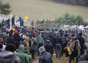 Migrantes param na cerca de arame farpado. Foto Leonid Scheglov